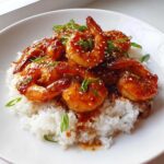 Plump shrimp coated in a glossy, sticky sweet chili sauce served over white rice, garnished with sesame seeds.