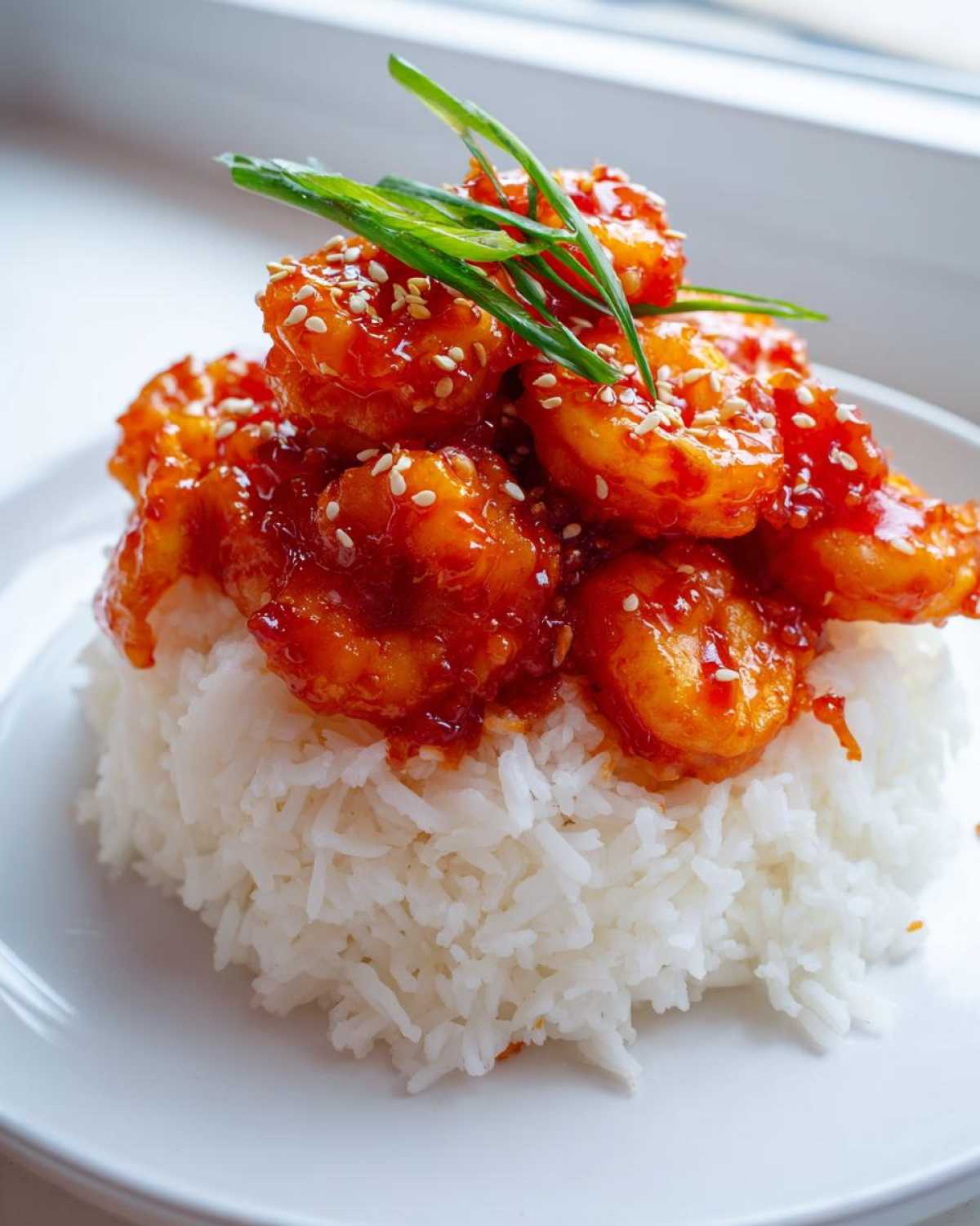 Close-up of Sticky Sweet Chili Shrimp glazed in a vibrant red sauce, served atop a mound of fluffy white rice and garnished with sesame seeds and green onions.