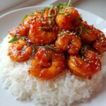 Plump shrimp coated in a glossy, orange-red sauce piled high on a bed of fluffy white rice, garnished with sesame seeds and green onions, showcasing Sticky Sweet Chili Shrimp.