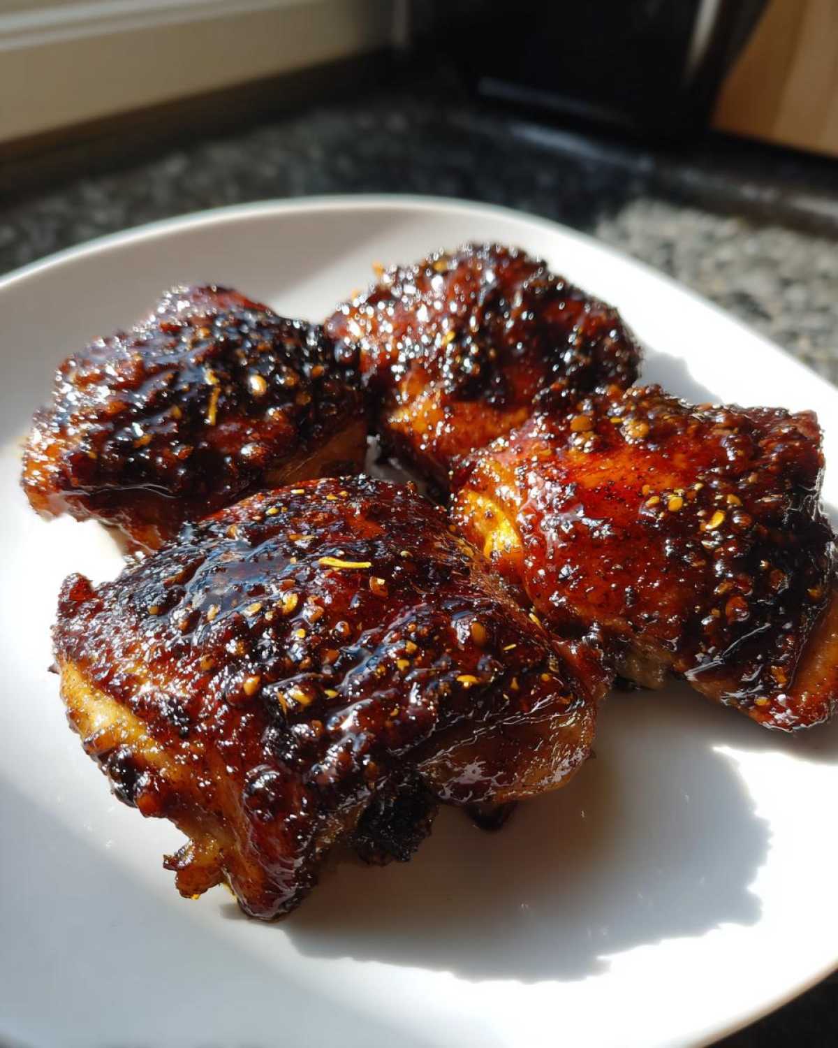 Four pieces of glazed Sticky Teriyaki Chicken Thighs with Garlic, glistening under bright light on a white plate.