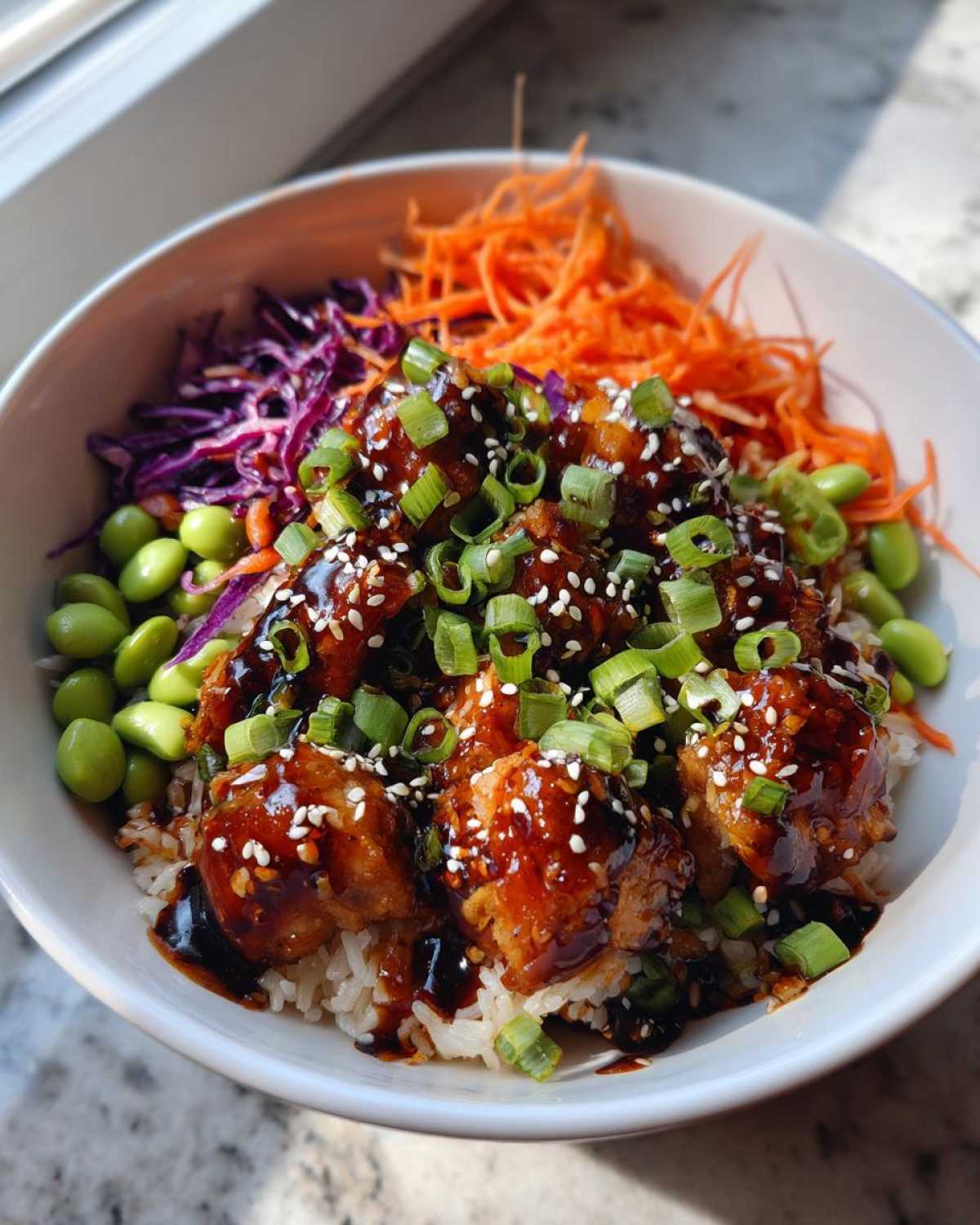 A close-up of a Sweet Chili Chicken Bowl with rice, topped with glazed chicken, shredded carrots, red cabbage, edamame, and green onions.