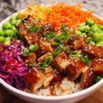 Close-up of a Sweet Chili Chicken Bowl featuring glazed chicken pieces over rice, topped with edamame, shredded carrots, and red cabbage.