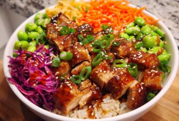 Close-up of a Sweet Chili Chicken Bowl featuring glazed chicken pieces over rice, topped with edamame, shredded carrots, and red cabbage.