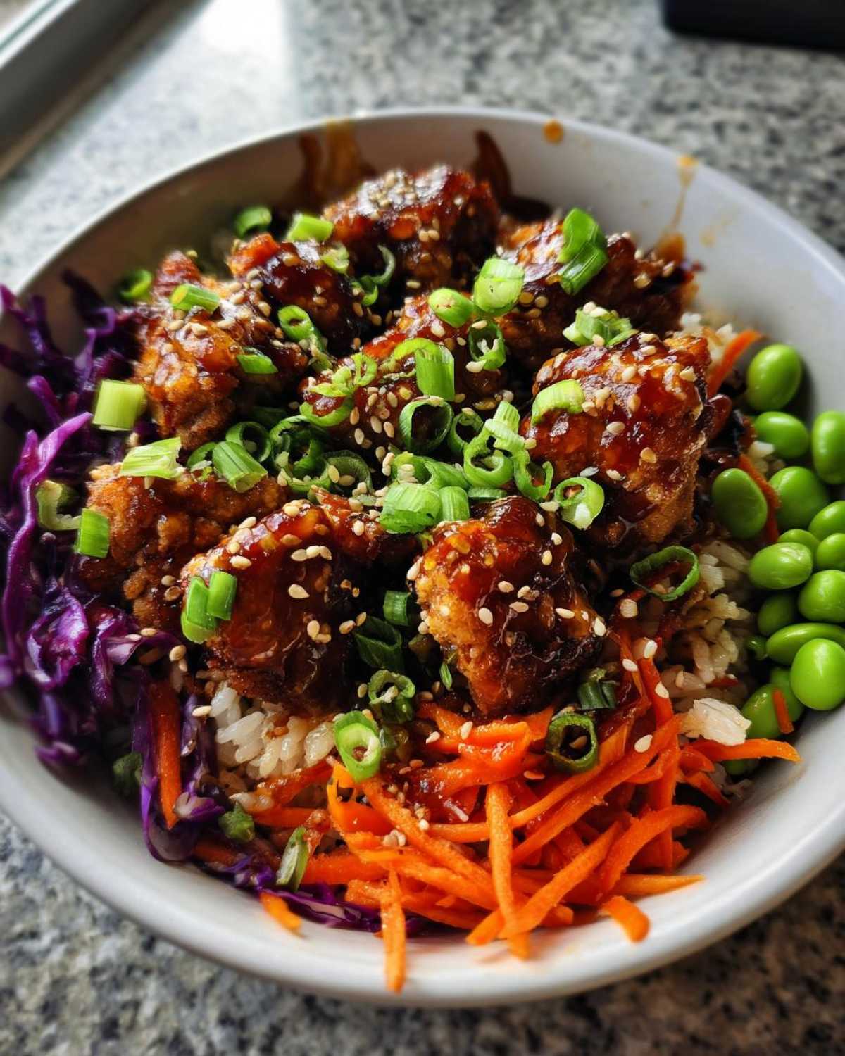 Close-up of a Sweet Chili Chicken Bowl featuring glazed chicken, shredded carrots, purple cabbage, edamame, and green onions.