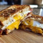 Cross-section of a Bacon Cheddar & Fried Egg Sandwich showing runny yolk, melted cheese, and crispy bacon on toasted bread.