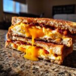A stacked Bacon Cheddar & Fried Egg Sandwich with bright yellow yolk dripping down onto a granite counter.