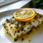 Close-up of a flaky, white piece of Baked Cod topped with herbs and a caramelized lemon slice.