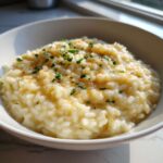 A close-up of creamy Parmesan Risotto topped with fresh parsley and a sprinkle of spice in a white bowl.