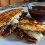 Two halves of a crispy quesadilla filled with shredded beef and melted cheese, served with a side of dark red consommé for dipping, typical of Crockpot Birria Tacos.