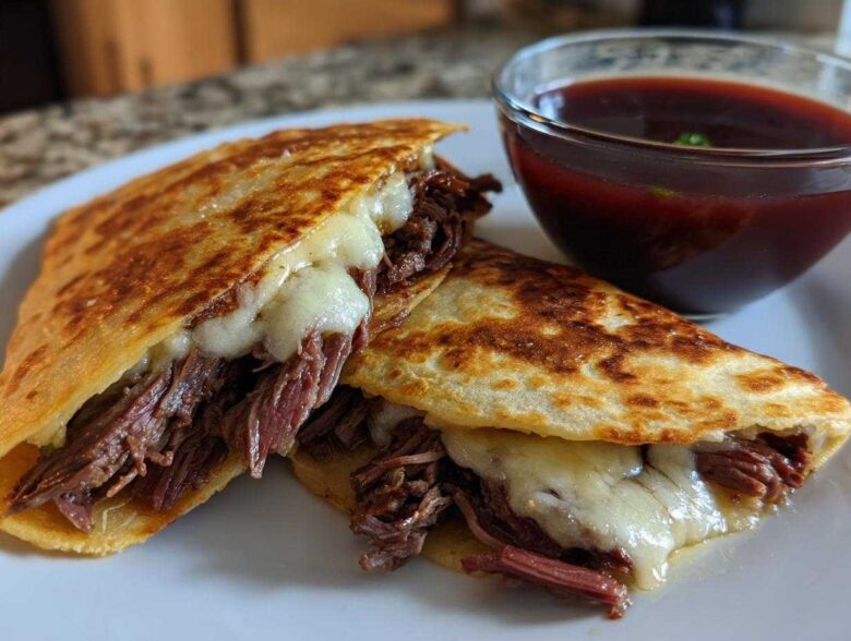 Two halves of a crispy quesadilla filled with shredded beef and melted cheese, served with a side of dark red consommé for dipping, typical of Crockpot Birria Tacos.