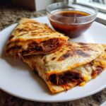 Two halves of a cheesy quesadilla filled with shredded beef, served next to a small bowl of consommé for dipping, perfect Crockpot Birria Tacos.