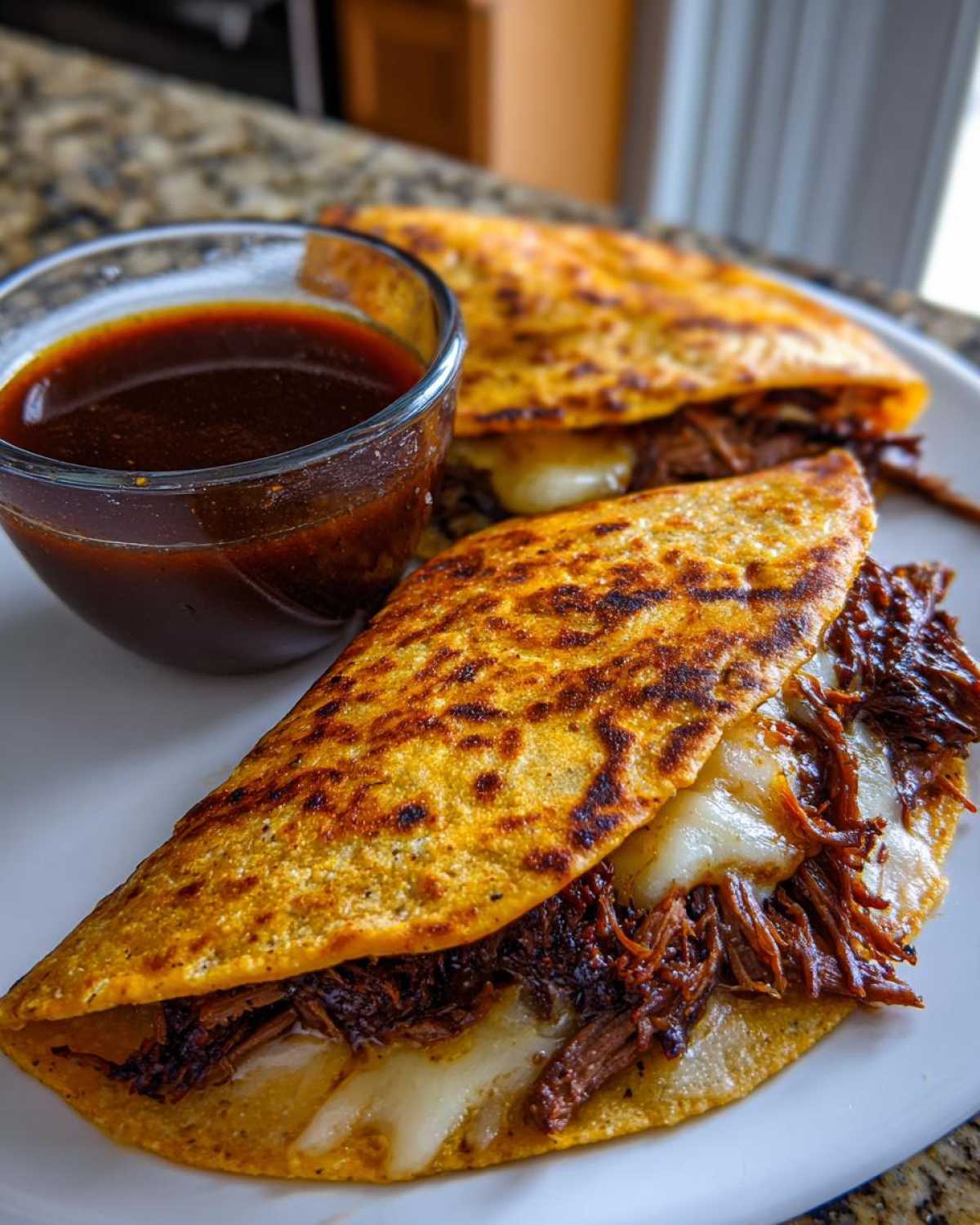Two crispy, cheesy Crockpot Birria Tacos served on a white plate next to a small bowl of dark consommé for dipping.