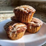 A stack of three moist Vanilla Chai Muffins topped with a thick white glaze, sitting on a white plate.