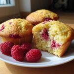 Three golden Lemon Raspberry Muffins on a white plate, one broken open to show the moist interior and bright raspberries.