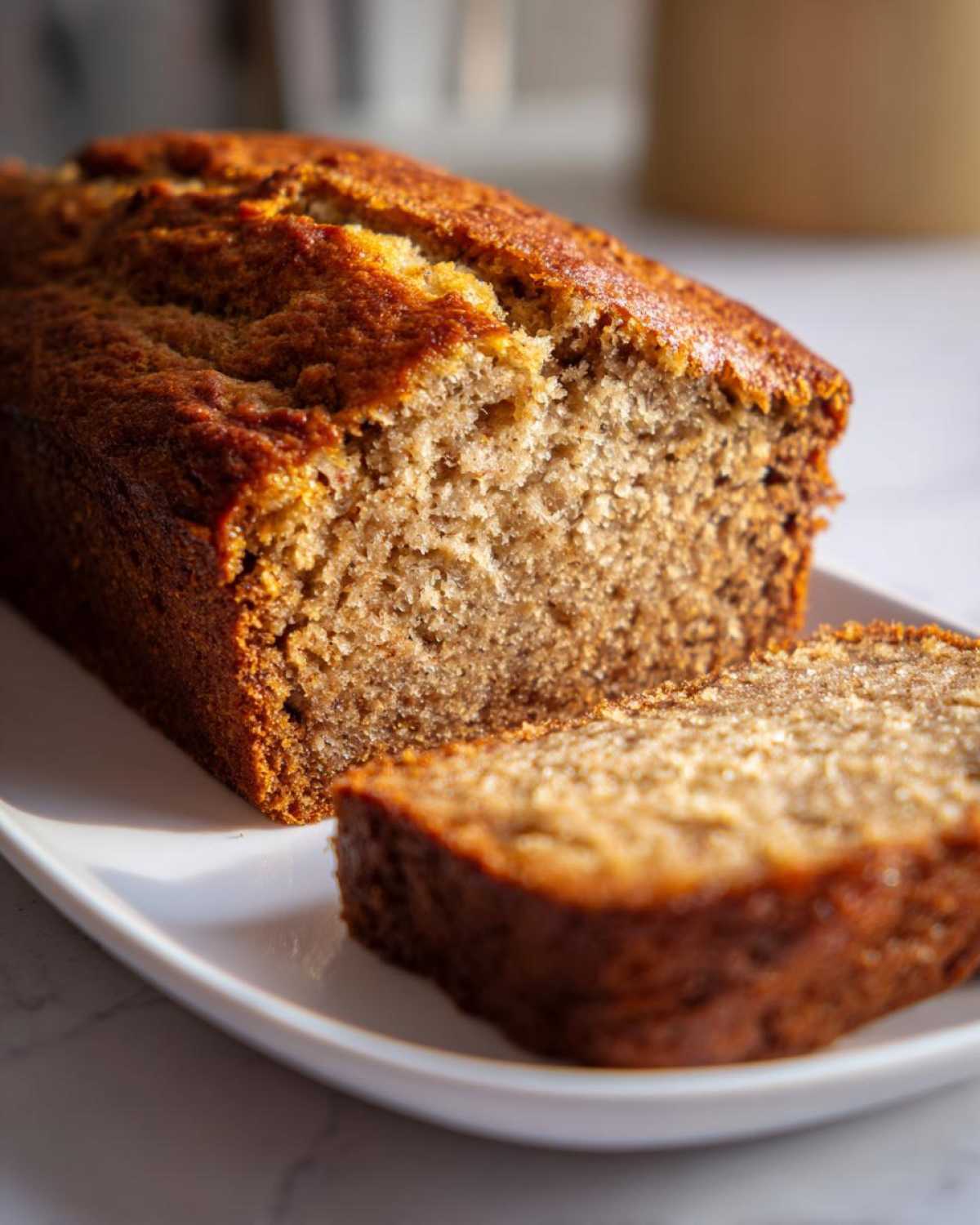 A golden brown loaf of moist Banana Bread, partially sliced on a white platter.