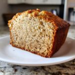 A thick slice of moist, golden-brown Banana Bread resting on a white plate.