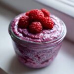 A close-up view of vibrant purple Raspberry Chia Seed Pudding topped with whole fresh raspberries in a glass jar.