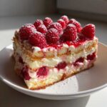 A close-up of a layered slice of Raspberry Tiramisu featuring ladyfingers, cream, and fresh raspberries, dusted with powdered sugar.
