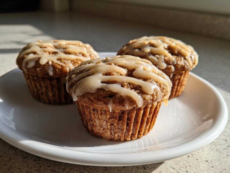 Three moist Vanilla Chai Muffins topped with a creamy glaze, resting on a white plate in natural sunlight.