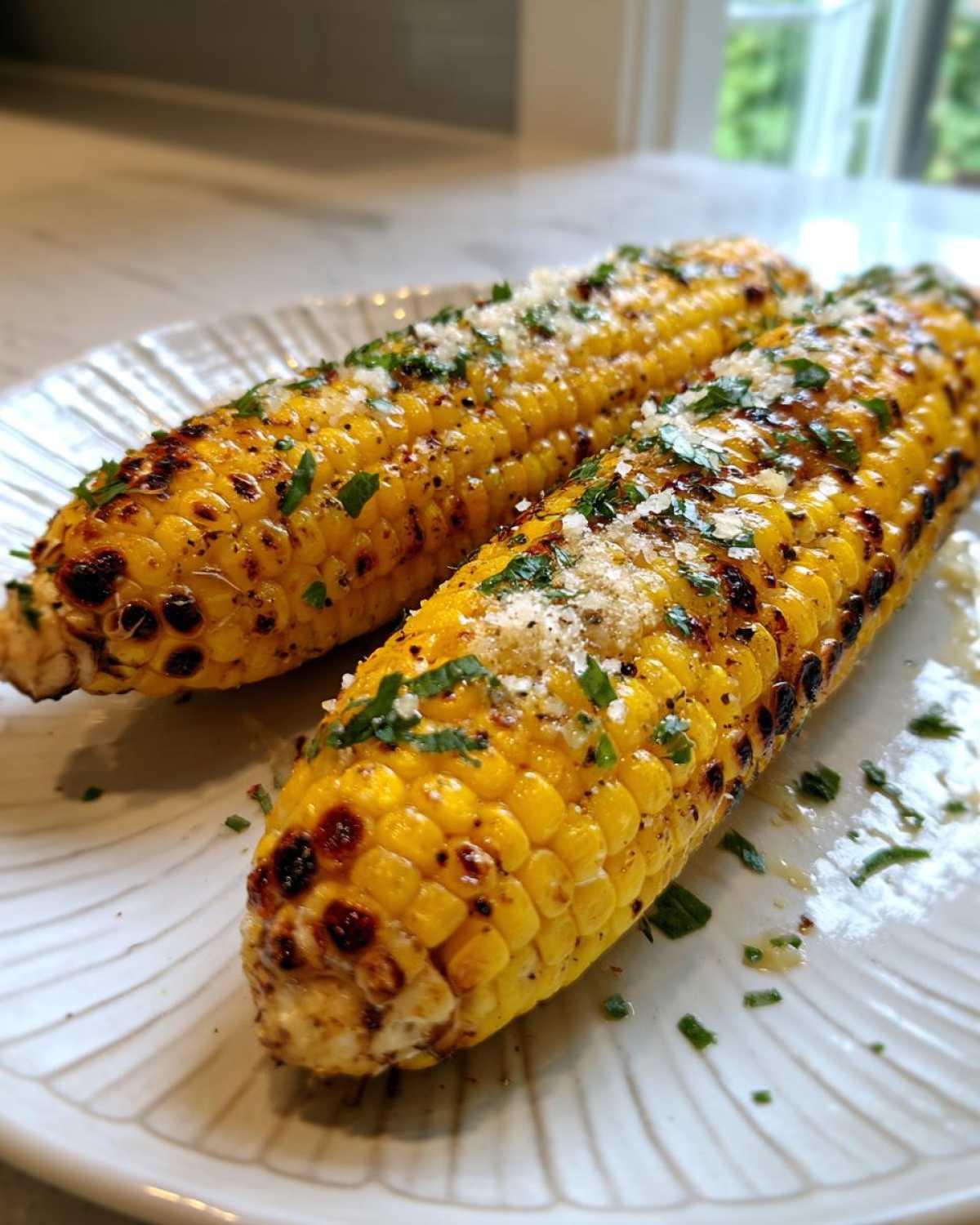Two ears of charred and seasoned Air Fryer Corn topped with grated cheese and fresh parsley on a white plate.