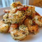 A close-up of cooked Air Fryer Garlic Parmesan Shrimp piled on a white plate, coated in herbs and crispy garlic.