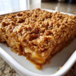 Close-up of a thick, gooey slice of homemade Apple Crisp with a crunchy oat topping in a white square baking dish.