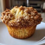 A single, golden Apple Cinnamon Crumb Muffin sits on a white plate, featuring a thick, sugary cinnamon crumb topping.