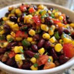 A close-up view of vibrant Cowboy Caviar Dip featuring black beans, yellow corn, red peppers, and green herbs in a white bowl.