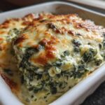Close-up of hot, baked creamy Spinach Dip topped with melted, golden-brown cheese in a white baking dish.