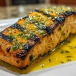 Close-up of a perfectly grilled salmon fillet generously topped with lemon butter sauce and fresh herbs.