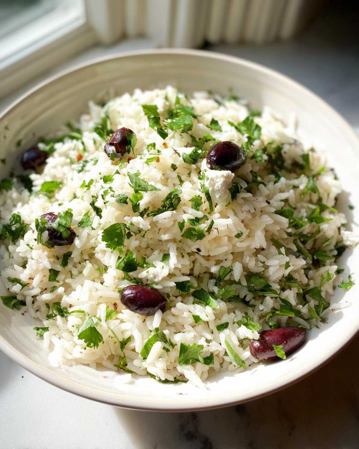 A bowl of fluffy white Mediterranean Rice mixed with fresh chopped parsley, Kalamata olives, and feta cheese.