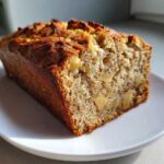 A moist slice of Apple Cinnamon Bread showing visible apple chunks, sitting on a white plate in bright sunlight.