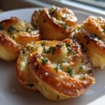 Four golden brown Puff Pastry Garlic Knots glistening with butter and topped with fresh chopped parsley on a white plate.