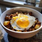 A close-up of Sausage and Potato Breakfast Bowls topped with a sunny-side-up egg and shredded cheese.