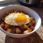 A white bowl filled with Sausage and Potato Breakfast Bowls, topped with a sunny-side-up egg and shredded cheese.
